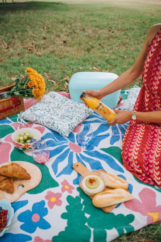 A person is pouring Limeñita at a picnic with food, flowers, and a pillow on a colorful blanket outside, perfect for breakfast.