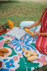 Colorful picnic setup on a blanket with Limeñita drink, fresh croissants, and salad outdoors.